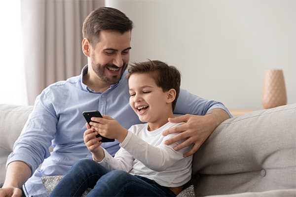 Young-father-embraces-little-son-family-sitting-on-couch-at-home-using-smart-phone-feels-happy